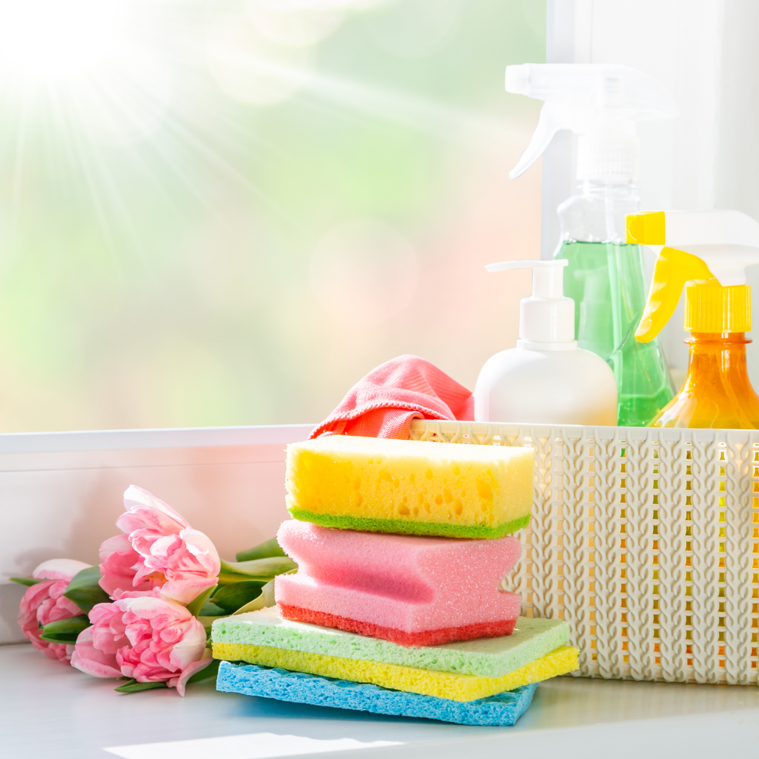 a basket of different cleaning supplies on a shelf by a window, with spray bottles and scrubbing sponges